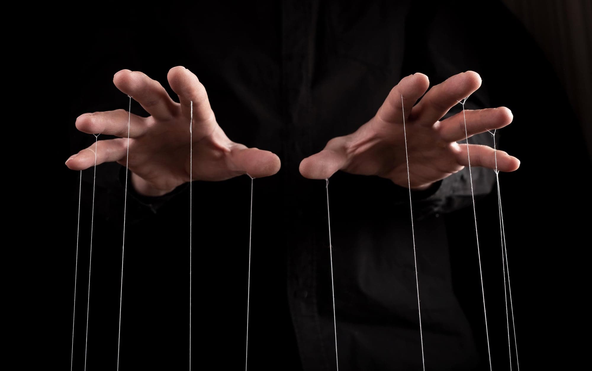 Close-up of hands manipulating puppet strings in darkness, symbolizing control, manipulation, and human power dynamics.